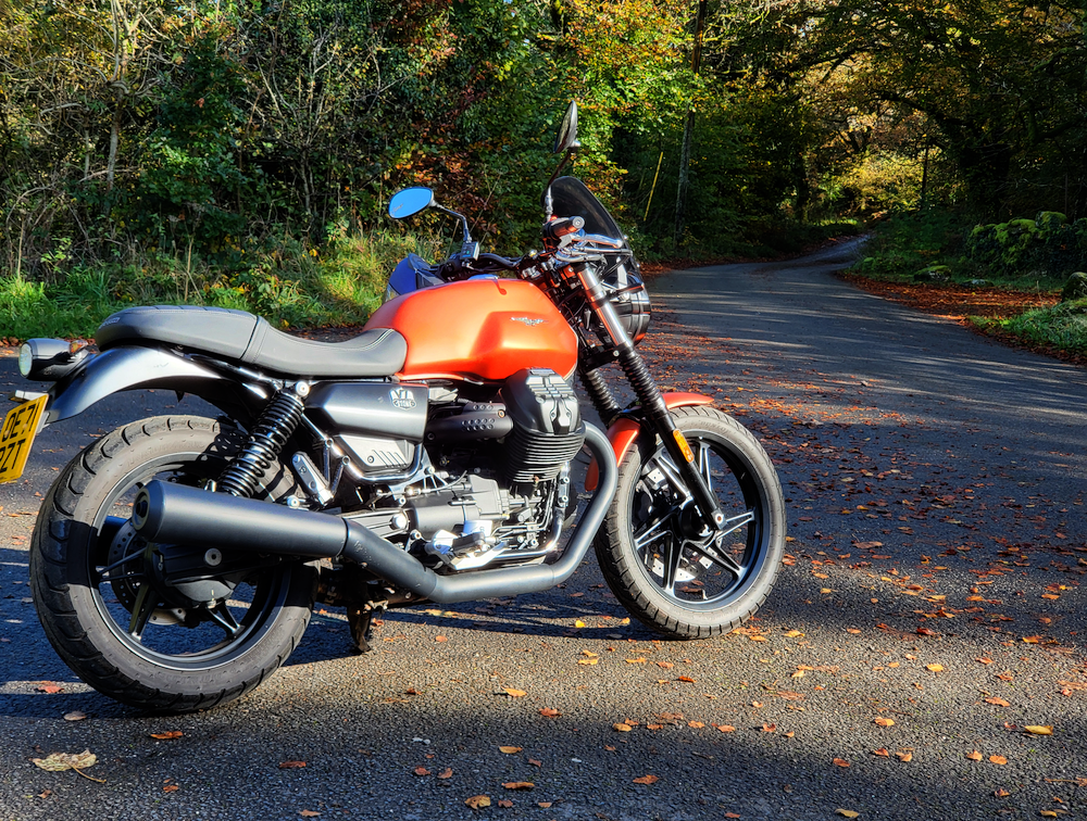 My V7, burnt orange. It’s on a grey tarmac road, pointing down it. it looks to be early autumn, on this country road